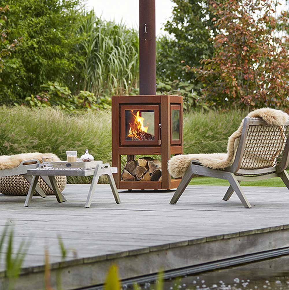 A modern cube-shaped Quaruba mobile outdoor log burner made of Corten steel with visible rust, placed on a patio next to a glass door leading to a garden. There is a fire burning inside the log burner and it's attached to a vertical chimney.