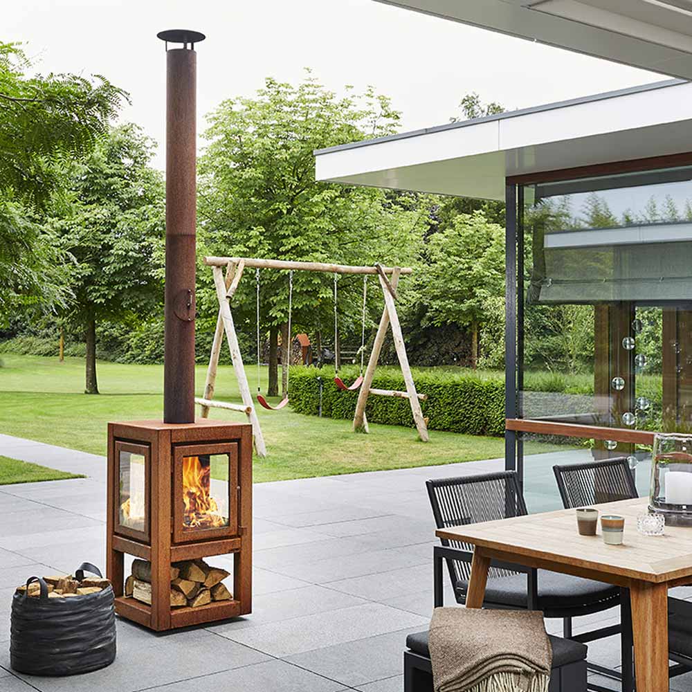 A modern cube-shaped Quaruba XXL mobile outdoor log burner made of Corten steel with visible rust, placed on a patio next to a glass door leading to a garden. There is a fire burning inside the log burner and it's attached to a vertical chimney.